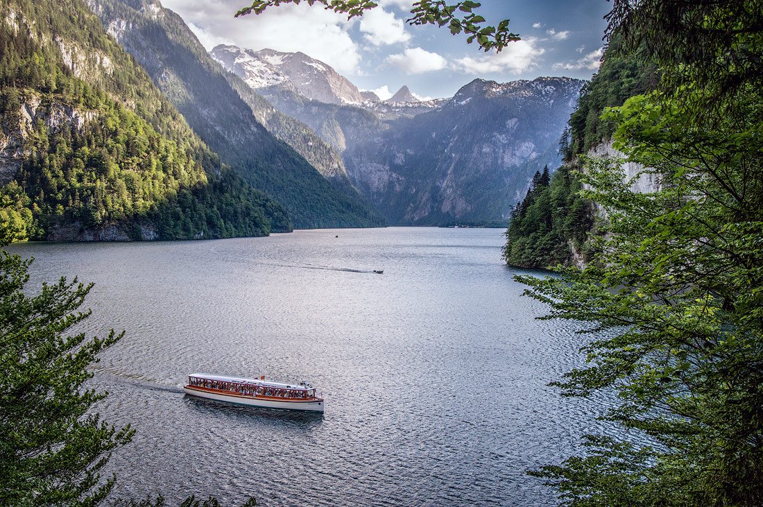 A boat cruising along the clear waters of Königssee, surrounded by towering cliffs and dense forests of the Bavarian Alps, under a partly cloudy sky