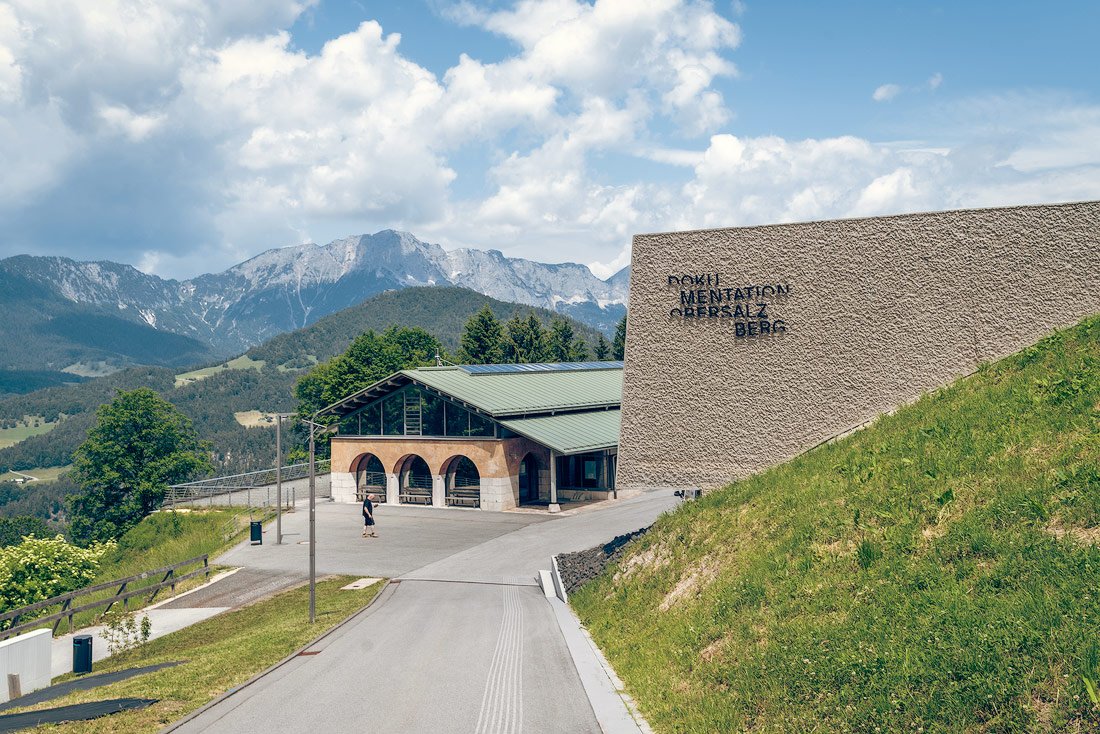 The Dokumentation Obersalzberg building on a sunny day, set against a backdrop of the Bavarian Alps, with its modern architecture and a pathway leading up to it.