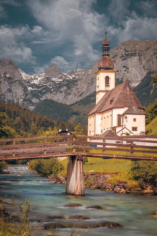 The Church of St. Sebastian in Ramsau with a wooden bridge crossing a clear mountain stream, set against the backdrop of the Bavarian Alps under a partly cloudy sky.