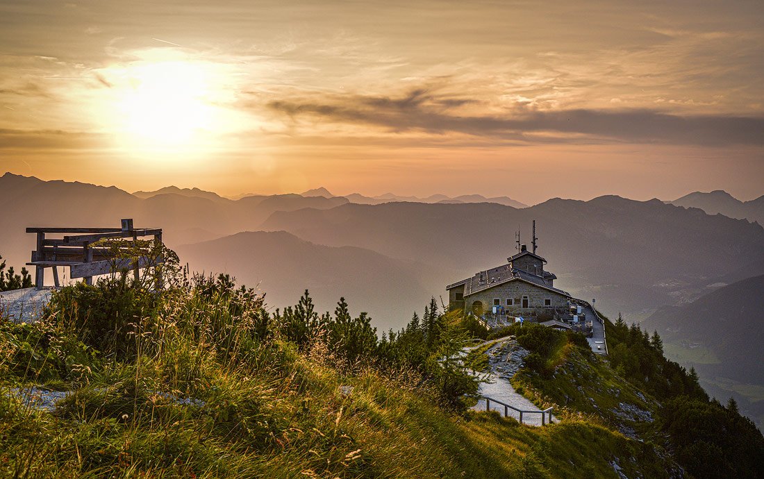 A view of the Eagle's Nest perched on a mountain peak during sunset, with a warm golden sky and layered mountain silhouettes in the background, and a wooden bench overlooking the scenery.