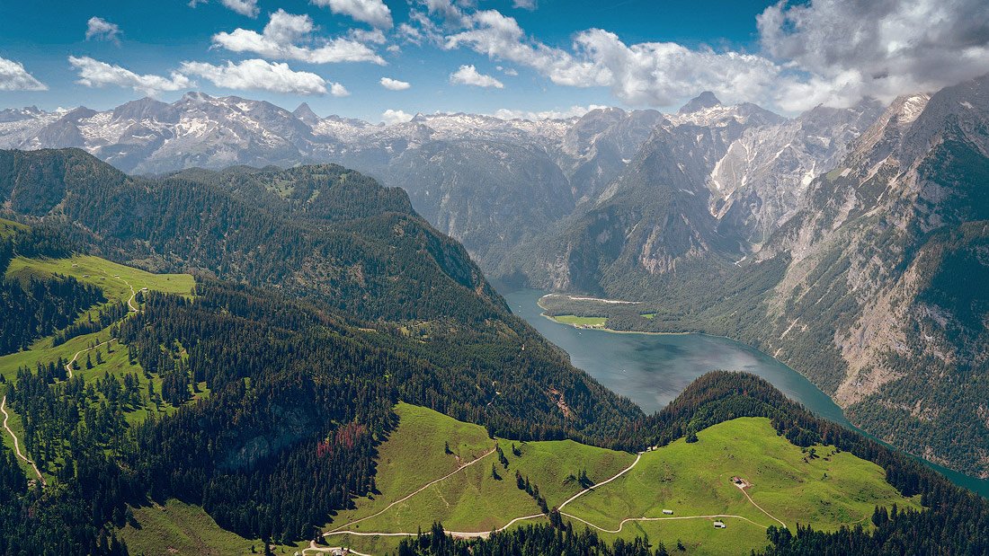 A panoramic view of the Bavarian Alps with snow-capped peaks in the background, a lush green valley, and the shimmering waters of Königssee nestled between the mountains under a clear blue sky.