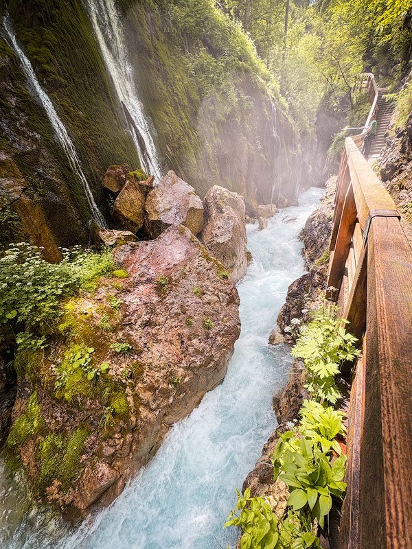 A narrow, rushing mountain stream flowing through a steep gorge in Wimbachklamm, surrounded by moss-covered rocks and lush greenery, with a wooden walkway alongside.